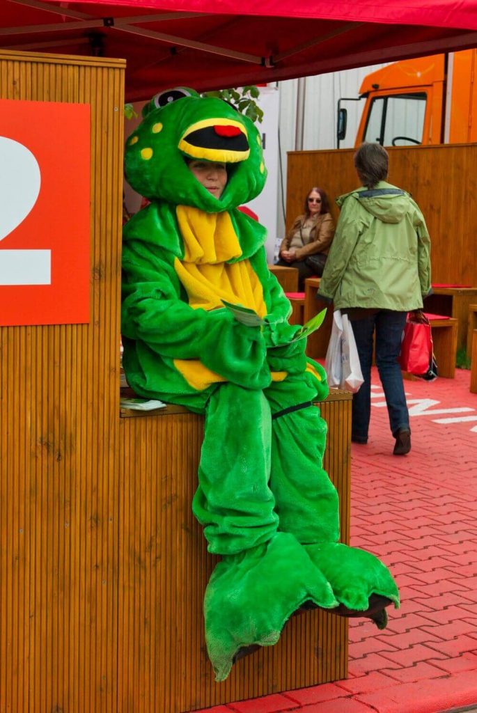Frau als Frosch verkleidet auf dem Mannheimer Maimarkt, sitz auf Theke und verteilt Flyer